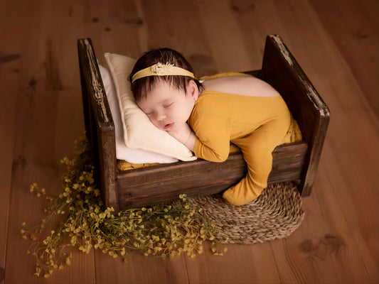 Newborn in a rustic wooden bed, yellow outfit, surrounded by soft yellow flowers, ideal newborn photography prop.